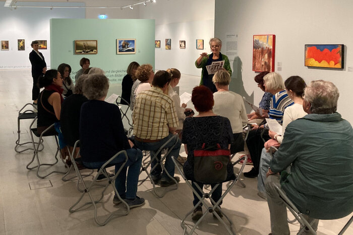 Eine Gruppe von Personen sitzt in einer Ausstellung, sie schauen auf eine Frau die etwas erklärt