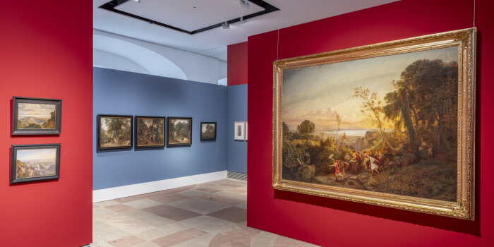 Ein Ausstellungsraum mit roten und blauen Wänden an den Gemälde hängen.