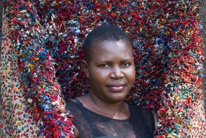 Hellen Nabukenya. Tuwaye – Let's talk eine dunkelhäutige Frau vor einem bunten Teppich