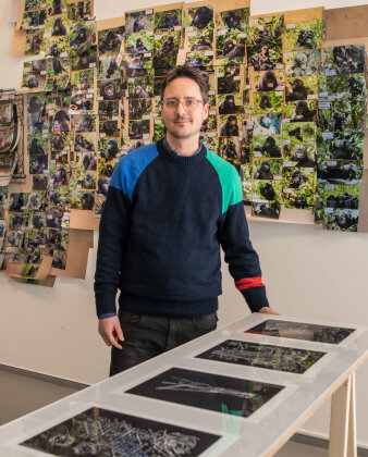 Ein Mann steht vor einer Wand mit Fotografien von Menschenaffen. Im Vordergrund eine Vitrine mit Drucken