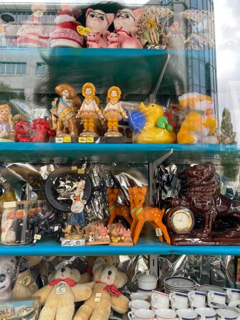Schaufenster mit verschiedenen Puppen, Figuren und Geschirr. Fotografierende Person spiegelt sich im Fensterglas