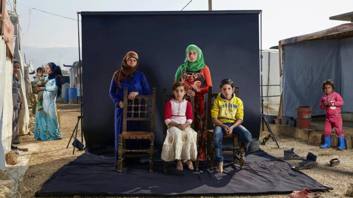„Khawle’s family“ im Flüchtlingslager Bekaa Valley im Libanon Vier Personen sitzen mittig vor einer schwarzen Leinwand drumherum Zelte und weiter Personen
