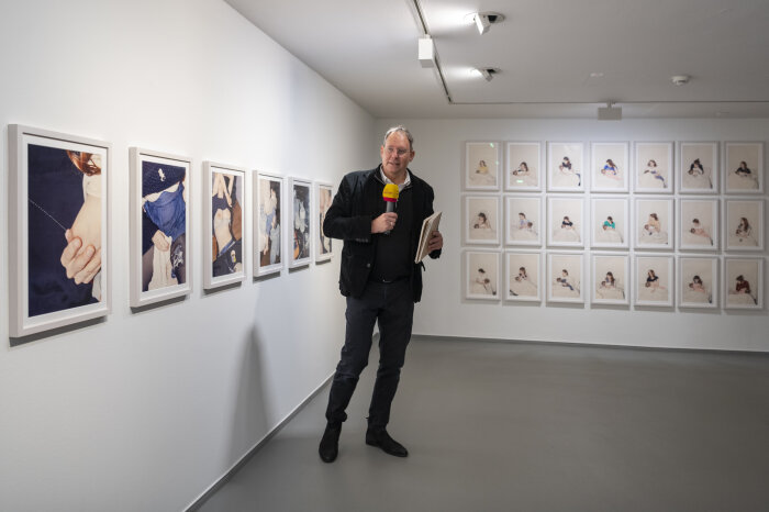 Kurator Ingo Taubhorn zum Pressegespräch Raum mit gerahmten Fotos an der Wand, in der Mitte ein stehender Mann mit Mikro in der Hand