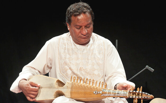 Musiker Ustad Ghulam Hussain (Rubab) Mann an einem gitarrenartigen Instrument vor dunklem Hintergrund.