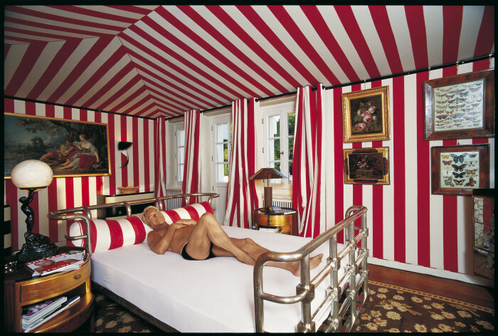 Aufnahme aus der Serie „Schlafzimmer“ (2002) Rot weiß gestreifter Raum mit Bildern in der Mitte ein Mann auf einem weißen Bett liegend