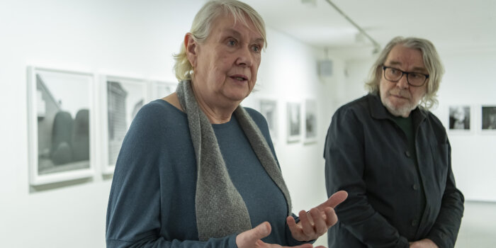 eine Frau mit grauen Haaren artikuliert mit den Händen, im Hintergrund steht ein Mann mit längerem grauen Haar