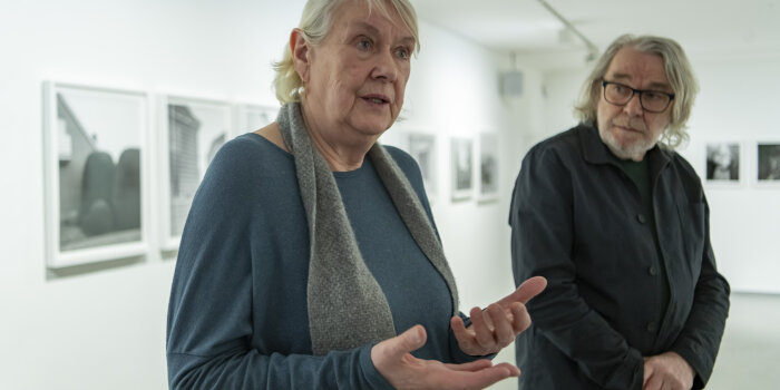 eine Frau mit grauen Haaren artikuliert mit den Händen, im Hintergrund steht ein Mann mit längerem grauen Haar