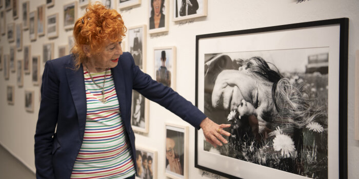 eine Frau mit roten Haaren zeigt erklärend auf eine Schwarz-Weiß-Fotografie des im Gras liegenden John Cage