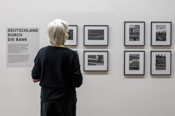 Eine dunkel angezogene Frau mit weißen Haaren schaut auf eine Wand mit Schwarz-Weiß-Fotografien