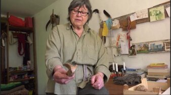 Foto: Still „Dorothea Prühl – Schmuck als Ornament und Skulptur“, D 2021 © Jochen Wisotzki Filmproduktion Eine ältere Frau steht in einem Raum mit Kunstwerken an der Wand und hält ein Objekt aus Holz