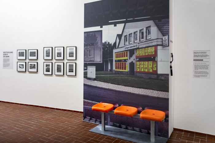 Ein großes Foto eines Gebäudes an einer Wand, davor drei Sitzgelegenheiten in Orange