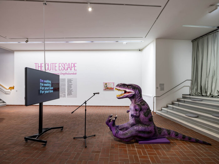 Eine Dinofigur mit Mikrofon in einem Raum sitzend vor einem Fernseher mit Schrift