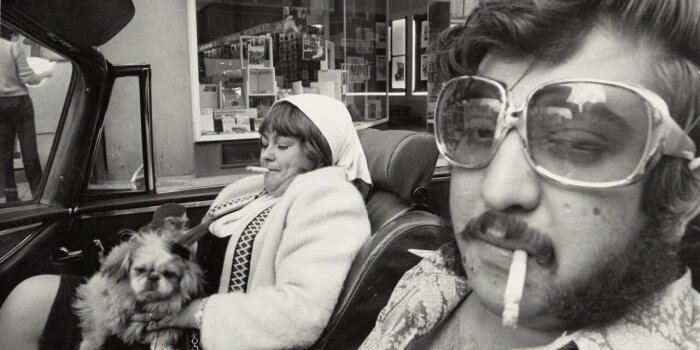 Mann und Frau in einem Cabriolet sitzend (Hamburg), 1971, aus der Serie „Was ist Schönheit?“, Silbergelatine-Abzug Ein schwarz weiß Foto eines rauchenden Mannes im Auto, daneben eine rauchende Frau mit einem Hund auf dem Schoß