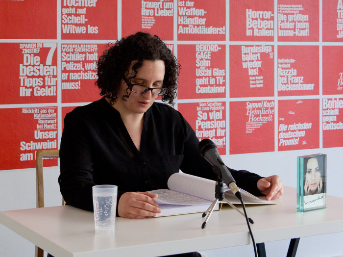 Künstlerin Anke Stiller bei einer Lesung. Im Hintergrund rote Plakate mit weißer Schrift.