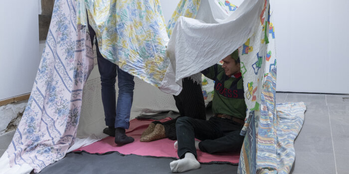 drei Personen sitzen, knien und stehen unter mehreren zusammengenähten Decken