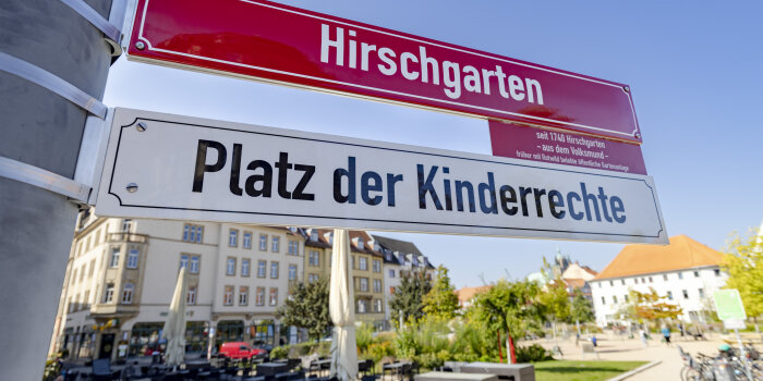 rotes Straßenschild "Hirschgarten" mit weißem Zuschatzschild "Platz der Kinderrechte"