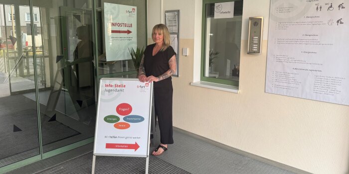 Eine Frau mit langen blonden Haaren steht an einem Schild. Darauf steht: Infostelle Jugendamt. 