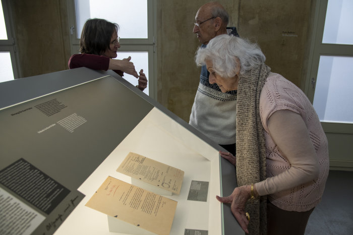 Éva Fahidi-Pusztai beim Besuch der Dauerausstellung "Techniker der Endlösung". Im Hintergrund: Dr. Annegret Schüle und Andor Andrási Eine Frau steht vor einer Vitrine und liest ein Dokument.