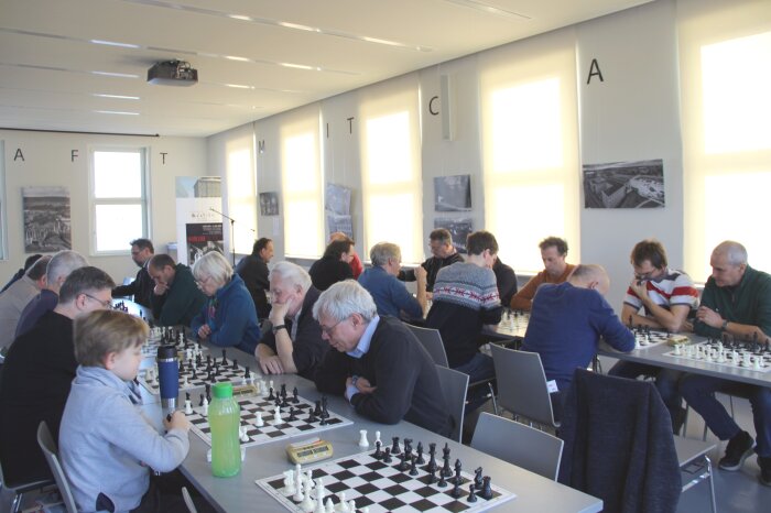 6. Königsgambit-Gedenkturnier, 2019 Farboto. Mehrere Personen sitzen an zwei langen Tischen und spielen Schach.