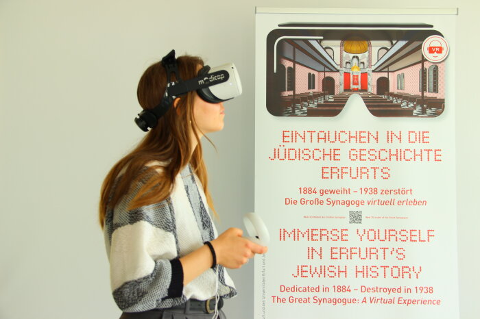Seminar mit VR Brille junge Frau mit VR Brille auf dem Kopf daneben ein Plakat mit der Aufschrift Eintauchen in die jüdische Geschichte