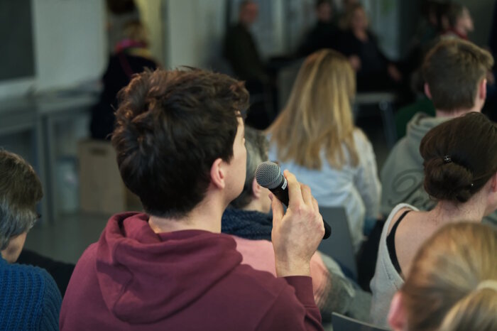 Besucher im Erinnerungsort Mehrere Personen von hinten und eine Poerson hat ein Mikrofon in der Hand
