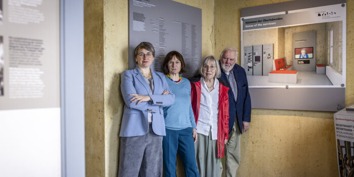 vier Personen stehen in einem Ausstellungsraum, an einer Wand hängt eine ausgedruckte Visualisierung eines Raumes
