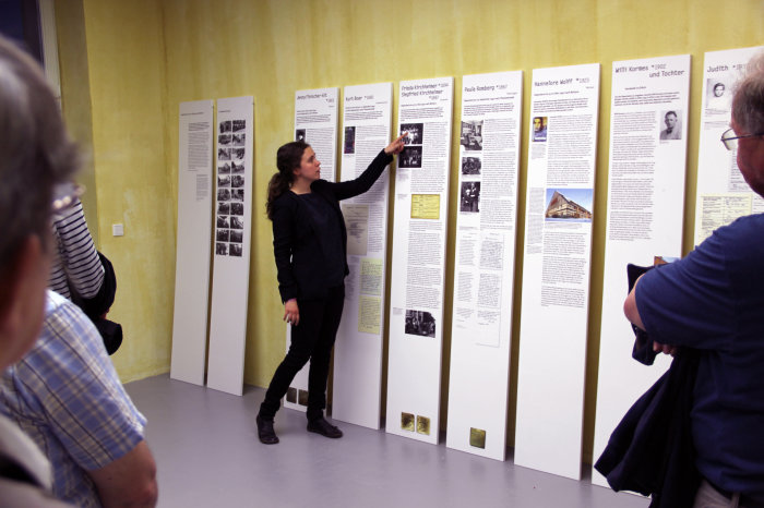 Biografische Kurzführungen in der Ausstellung "Jüdische Nachbarn" Farbfotografie, junge Frau steht vor einer Reihe bedruckter weißer Stelen, die an eine gelbe Wand gelehnt sind, sie zeigt auf ein Bild