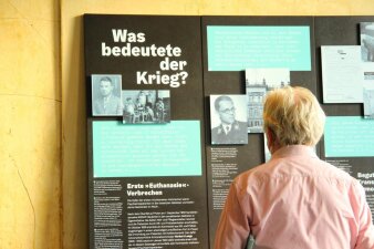 Person vor einer schzwarzen Wand mit Textfeldern und Bildern mit der Aufschrift Was bedeutet der Krieg?