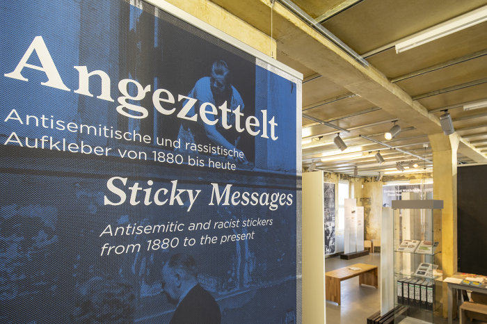Blick in die Ausstellung „Angezettelt. Antisemitische und rassistische Aufkleber von 1880 bis heute“ Eine Tafel und Ausstellungsvitrinen