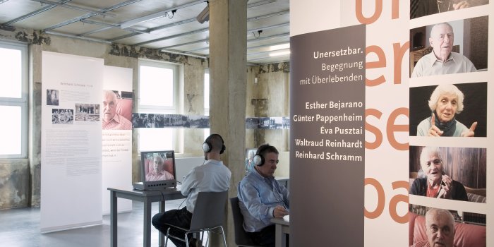 Foto: Blick in die Ausstellung "Un-er-setz-bar. Begegnung mit Überlebenden" © Stadtverwaltung Erfurt Blick in den Raum mit Tafeln. Besucher benutzen Multimediageräte