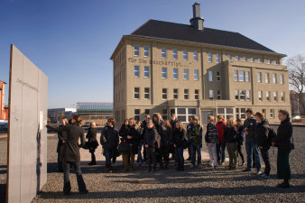 Im Vordergrund eine Gruppe von Jugendlichen, die zu einer weibliche Person links im Bild vor einer grauen Stele schaut.Im Hintergrund Gebäude mit Spitzdach