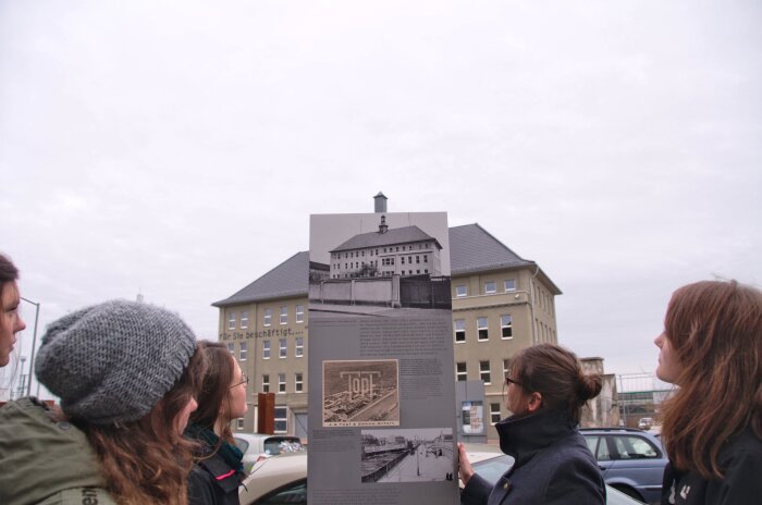 Eine Gruppe schaut auf eine Informationsstehle. Im Hintergrund das Gebäude des Erinnerungsortes Topf & Söhne.