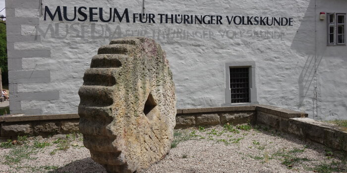 Foto: Waidmühlstein © Stadtverwaltung Erfurt/ Museum für Thüringer Volkskunde großes Steinrad vor Hauswand mit der Schrift Museum für Thüringer Volkskunde